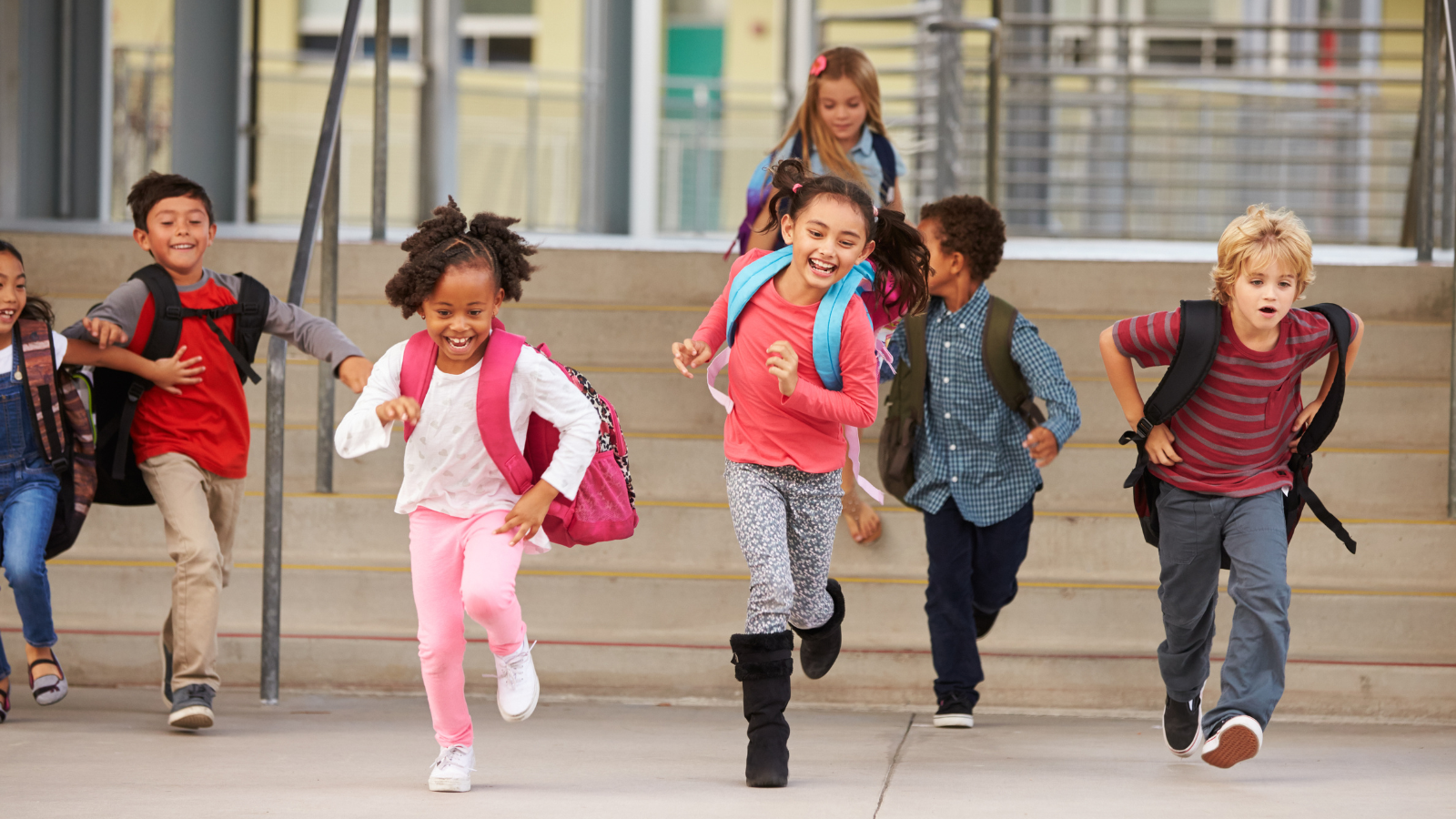 Happy children running down an outdoor school staircase, with school doors in the background.